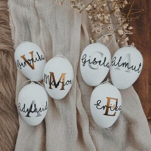 Op de afbeelding: Vijf witte paaseieren met gouden en zilveren letters. De eieren zijn versierd met de namen "Marion", "Erika", "Gisela Almut", "Lydia Heid" en "Friederike".