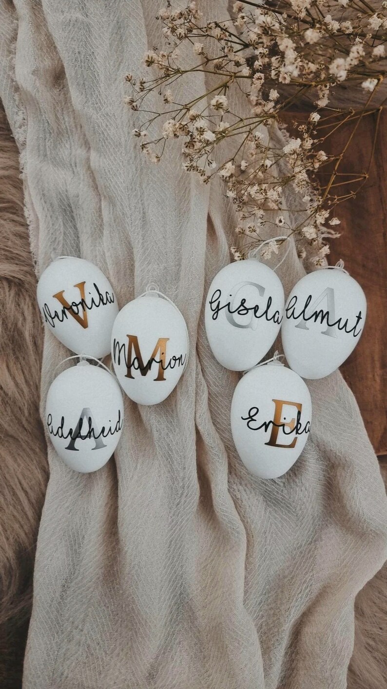 Op de afbeelding: Vijf witte paaseieren met gouden en zilveren letters. De eieren zijn versierd met de namen "Marion", "Erika", "Gisela Almut", "Lydia Heid" en "Friederike".