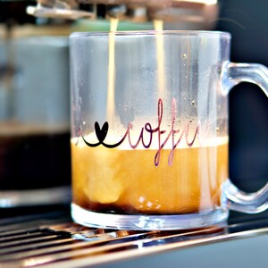 Puede incluir: Una taza de vidrio transparente con un coraz&oacute;n negro y la palabra "coffee" escrita en cursiva en el lateral. La taza se est&aacute; llenando de caf&eacute; de una cafetera.