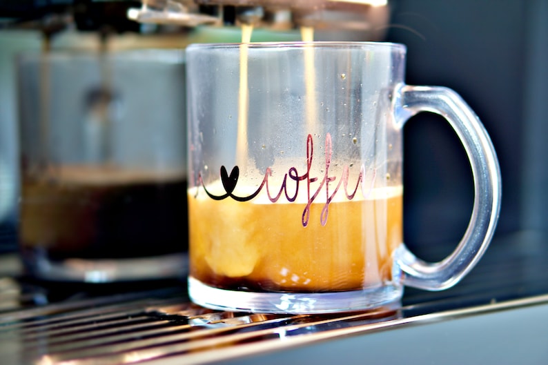 Puede incluir: Una taza de vidrio transparente con un coraz&oacute;n negro y la palabra "coffee" escrita en cursiva en el lateral. La taza se est&aacute; llenando de caf&eacute; de una cafetera.