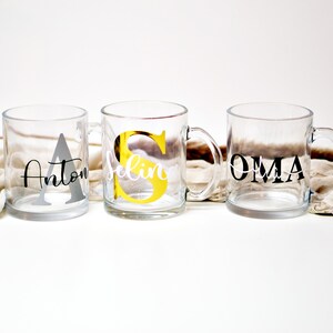 Puede incluir: Tres tazas de vidrio transparente con nombres y letras iniciales personalizadas. Las tazas est&aacute;n sobre una superficie blanca con una tela beige en el fondo. Los nombres en las tazas son Anton, Selin y Oma.