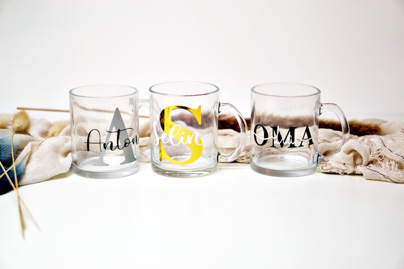 Puede incluir: Tres tazas de vidrio transparente con nombres y letras iniciales personalizadas. Las tazas est&aacute;n sobre una superficie blanca con una tela beige en el fondo. Los nombres en las tazas son Anton, Selin y Oma.