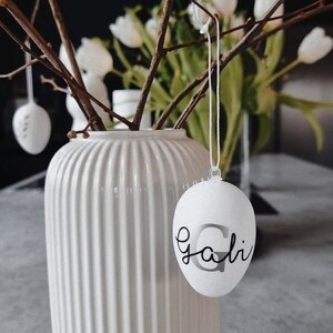 Op de afbeelding: Witte keramische vaas met een geribbelde textuur, gevuld met kale takken en witte bloemen. Een wit ei met het woord "Gali" erop hangt aan een tak.