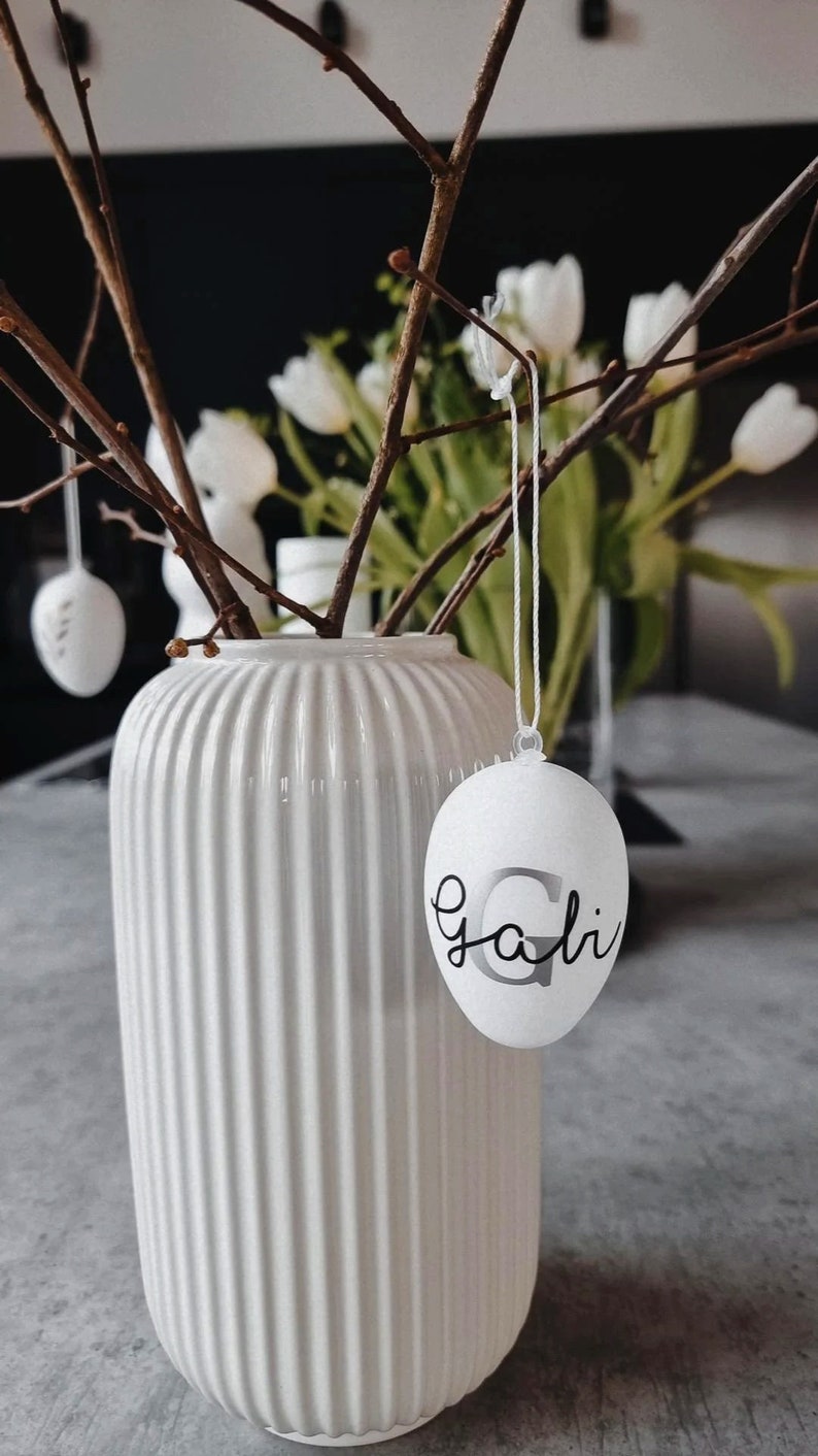Op de afbeelding: Witte keramische vaas met een geribbelde textuur, gevuld met kale takken en witte bloemen. Een wit ei met het woord "Gali" erop hangt aan een tak.