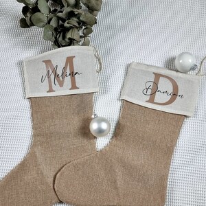 K&ouml;nnte beinhalten: Zwei personalisierte Weihnachtsstr&uuml;mpfe aus Jute mit den Namen "Melina" und "Damian", die in brauner Schrift auf der Oberseite jedes Strumpfes stehen.