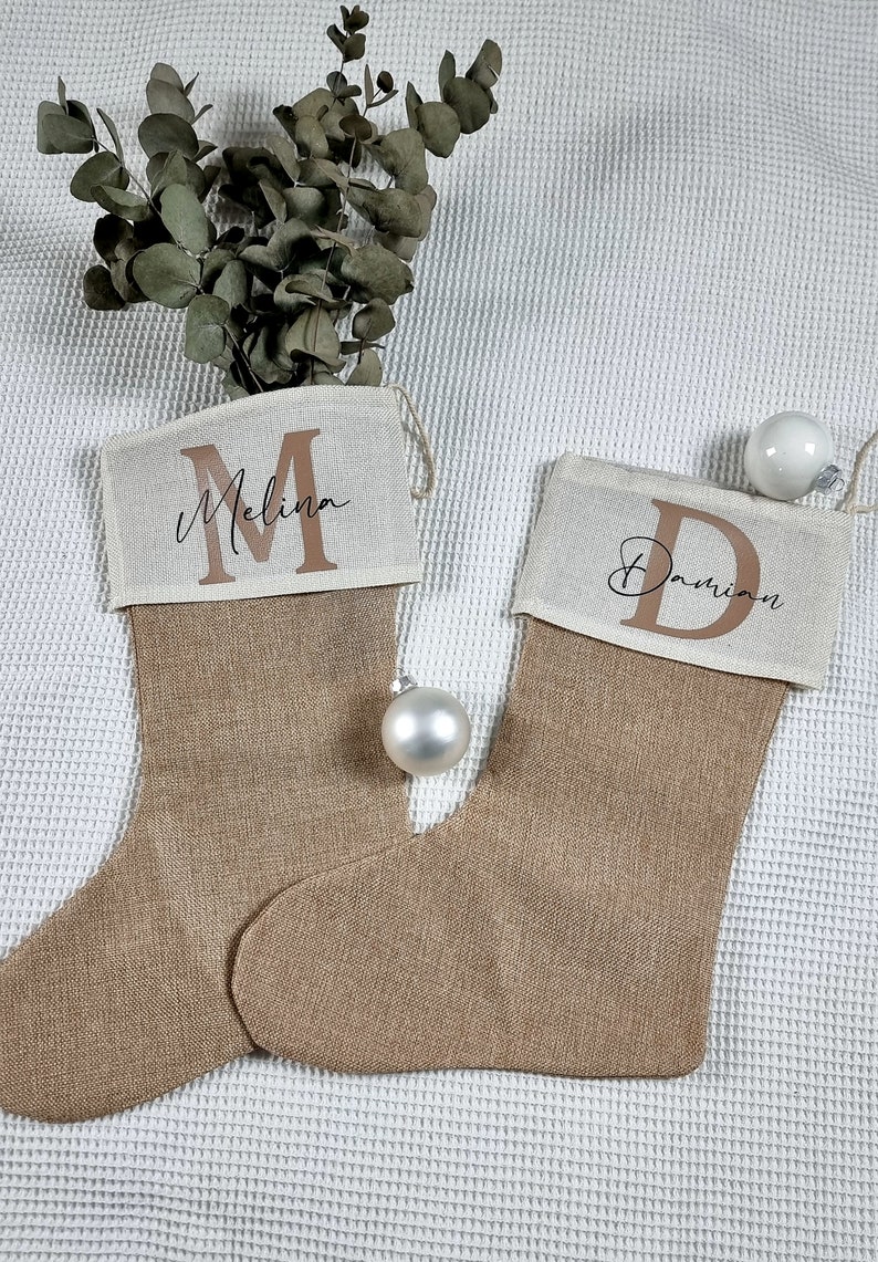 K&ouml;nnte beinhalten: Zwei personalisierte Weihnachtsstr&uuml;mpfe aus Jute mit den Namen "Melina" und "Damian", die in brauner Schrift auf der Oberseite jedes Strumpfes stehen.