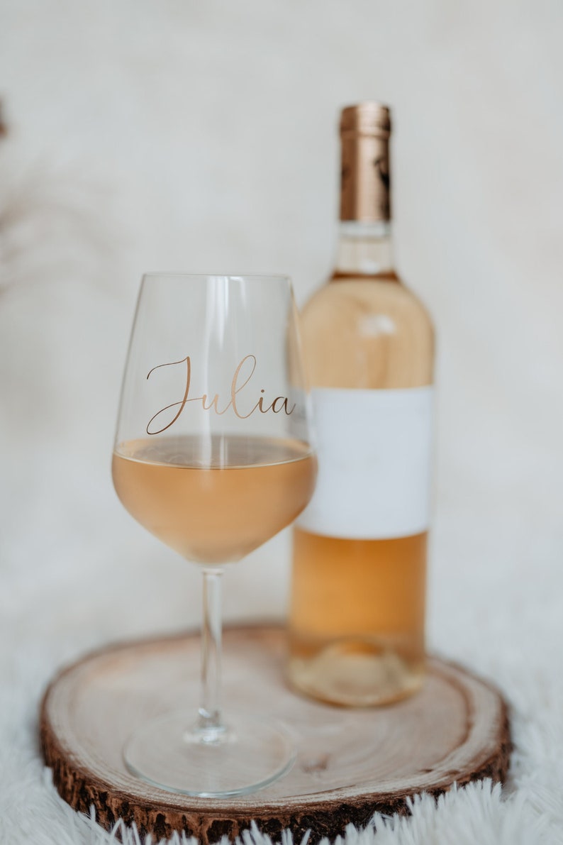 May include: A glass of white wine with the name "Julia" written on it, next to a bottle of white wine on a wooden slice.