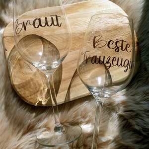 May include: Two clear wine glasses with gold lettering. The glass on the left says "Braut" and the glass on the right says "Beste Trauzeugin".