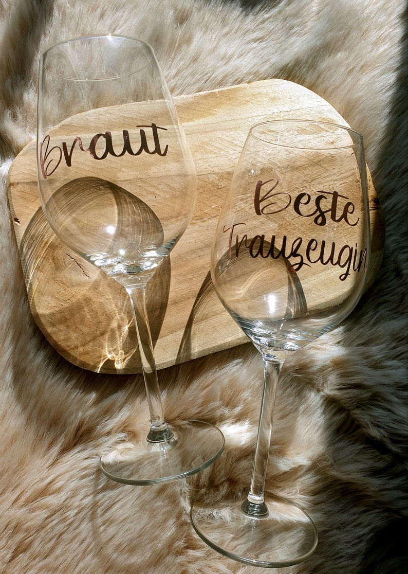 May include: Two clear wine glasses with gold lettering. The glass on the left says "Braut" and the glass on the right says "Beste Trauzeugin".