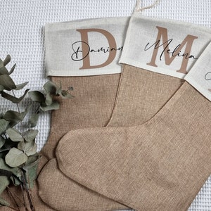 K&ouml;nnte beinhalten: Zwei braune Weihnachtsstr&uuml;mpfe aus grobem Leinen mit wei&szlig;en Stoffoberteilen. Die Str&uuml;mpfe sind mit den Namen "Damian" und "Melina" in brauner Schrift personalisiert.