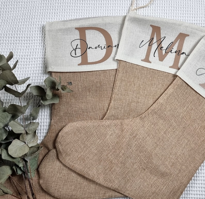 K&ouml;nnte beinhalten: Zwei braune Weihnachtsstr&uuml;mpfe aus grobem Leinen mit wei&szlig;en Stoffoberteilen. Die Str&uuml;mpfe sind mit den Namen "Damian" und "Melina" in brauner Schrift personalisiert.