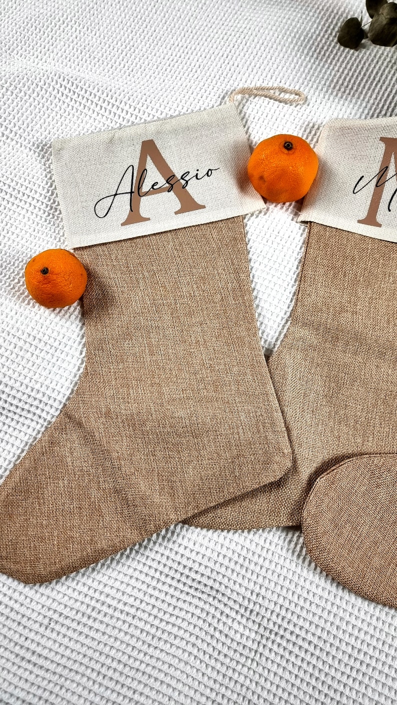 K&ouml;nnte beinhalten: Zwei braune Weihnachtsstr&uuml;mpfe aus grobem Leinen mit wei&szlig;en Stoffoberteilen. Die Str&uuml;mpfe tragen die Namen "Alessio" und "Mia", die in schwarzer Kursivschrift darauf geschrieben sind. Die Str&uuml;mpfe liegen auf einer wei&szlig;en, strukturierten Oberfl&auml;che mit zwei kleinen Orangen.