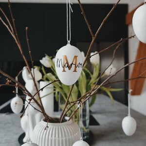 Op de afbeelding: Witte paaseieren met gouden letters die aan een tak hangen. De eieren zijn versierd met de namen "Marion", "Dennis" en "Gashi".