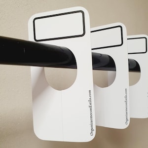 Può includere: Tre divisori per armadio bianchi sospesi con un foro rotondo al centro e uno spazio rettangolare in alto per scrivere. I divisori hanno il testo "OrganizarmneconEstilo.com" stampato in basso.