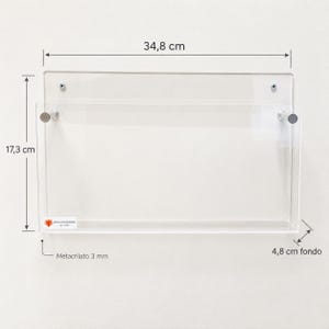 May include: A clear acrylic wall-mounted document holder measuring 34.8 cm wide, 17.3 cm high, and 4.8 cm deep. The unit is constructed from 3 mm thick acrylic and features two silver-toned metal standoffs for mounting, with a small logo label.