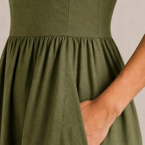 以下が含まれることがあります： 垂直の縫い目が入ったフィット感のあるボディスと、ギャザーが寄せられたウエストラインから広がるフルスカートが特徴のオリーブグリーンのドレス。スカートのサイドシームには機能的なポケットが組み込まれています。