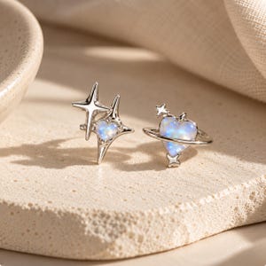 Può includere: Un paio di orecchini color argento con motivi celestiali. Un orecchino presenta una stella a quattro punte con un piccolo cuore iridescente al centro, mentre l'altro mostra una pietra iridescente a forma di cuore circondata da un anello con due piccole stelle.