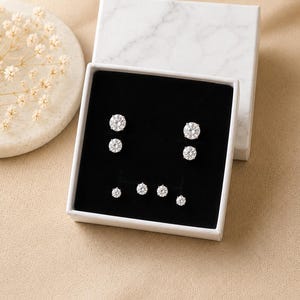 May include: A set of three pairs of round, clear crystal stud earrings displayed in a white box with a black interior. The box is open, revealing the earrings in varying sizes. The box sits on a beige surface, with a decorative white plate and dried flowers in the background.