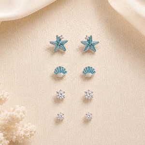 Può includere: Un set di quattro paia di orecchini a lobo disposti in una colonna verticale. La coppia superiore presenta stelle marine blu con piccole pietre chiare, la seconda coppia mostra forme di conchiglie blu e le due coppie inferiori sono pietre chiare rotonde incastonate.