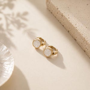 May include: A pair of gold-toned hoop earrings featuring a circular design with a smooth, white, iridescent inlay at the center. The earrings have a polished metallic finish and a hinged closure mechanism, presenting a clean and simple aesthetic.