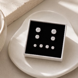 May include: A set of six round, clear, faceted stud earrings arranged in a square black display box. The earrings are organized in three pairs of varying sizes, featuring metallic prongs that secure each stone in a classic, circular setting.