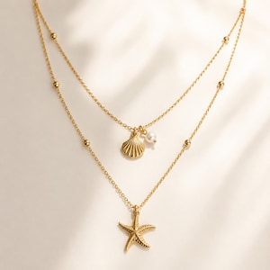 May include: A gold-toned double-layered necklace featuring a delicate chain with small beads. The upper layer holds a small seashell charm and a single white pearl, while the lower layer features a textured starfish pendant as the focal point.