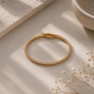 May include: A gold-toned tennis bracelet featuring a continuous row of small, clear, sparkling stones set in a metallic frame. The bracelet includes a secure, hinged clasp mechanism at the top, designed to hold the circular shape firmly in place.