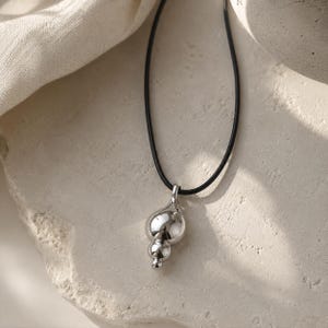 May include: A silver-toned pendant featuring three stacked, smooth, spherical shapes of varying sizes, suspended from a thin black cord. The metal surface is highly reflective, showing a polished finish that catches the light.
