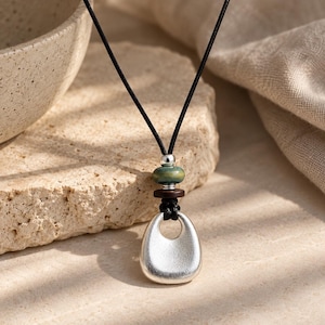 May include: A necklace featuring a black cord with a silver-toned pendant shaped like a rounded triangle with a central cutout. Above the pendant are a dark brown wooden disc, a small green ceramic bead, and a tiny silver-toned metal spacer bead.