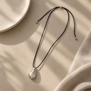 May include: A silver-toned teardrop pendant suspended on a thin black cord. The pendant has a smooth, reflective metallic surface. The black cord features two small silver-toned spherical beads at the ends, allowing for an adjustable length.