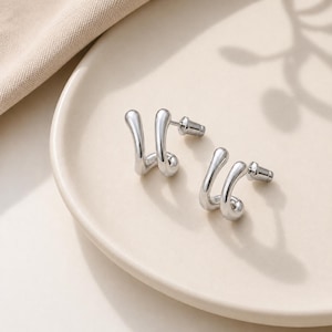 May include: A pair of silver-colored stud earrings with a unique, curved design. The earrings have a smooth, polished finish and are displayed on a light beige plate. The design features a flowing, abstract shape, adding a modern touch to the jewelry.