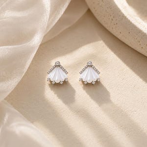 May include: A pair of silver-toned stud earrings shaped like a fan or shell. Each earring features a smooth, white, pearlescent center piece framed by a row of small, clear, sparkling stones along the top edge and at the base points.