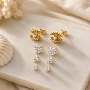 Puede incluir: Un conjunto de cinco pares de pendientes de botón en tono dorado. El par superior presenta un diseño de concha con una pequeña perla blanca. Los cuatro pares restantes son piedras redondas, transparentes y facetadas de tamaños graduados.