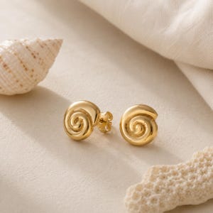 May include: A pair of gold stud earrings shaped like spiral seashells. The earrings feature a polished, smooth metallic finish with a distinct coiled design that mimics the natural form of a nautilus shell, complete with standard post backings.