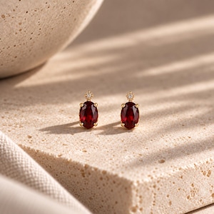 May include: A pair of gold stud earrings featuring oval-cut deep red garnet gemstones. Each earring is accented with a small, round, clear sparkling stone set in a star-shaped gold mount positioned directly above the primary red oval gemstone.