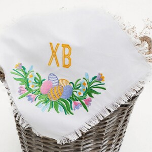Op de afbeelding: Witte geborduurde keukendoek met een gefranst randje. De borduurwerk toont een bloemmotief met kleurrijke paaseieren en de letters "XB".