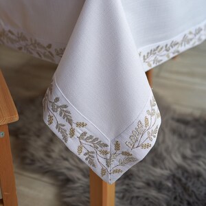 Puede incluir: Mantel de lino blanco con un borde bordado con hojas doradas. La mesa est&aacute; puesta con servilletas, platos y cubiertos a juego, creando un ambiente elegante.