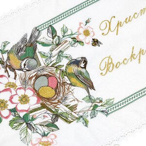 以下が含まれることがあります： 花と鳥のデザインが施された白い刺繍生地。刺繍には、2羽の青と黄色の鳥、卵のある巣、ピンクの花、そして金色の文字で「Христос Воскрес」という文字が描かれています。緑色の縁とレースが縁を飾っています。