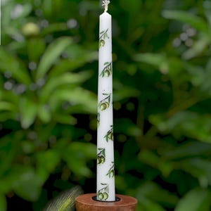 Op de afbeelding: Een witte kaars met een groen olijftakpatroon, geplaatst in een houten kandelaar. De kaars is aangestoken en heeft een wit label in de linkerbovenhoek met de tekst "Style 7". De achtergrond is een wazig groen gebladerte.
