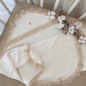 May include: White baby blanket with a cross embroidered on it. The blanket has a lace trim and is folded in half. The blanket is lying on a white crib mattress.