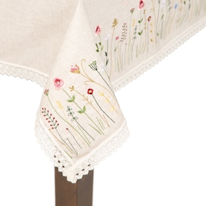May include: A white linen tablecloth with a delicate floral embroidery design. The tablecloth features a white lace trim and is shown draped over a wooden table.