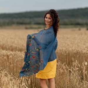 May include: A woman in a yellow dress and a blue floral scarf stands in a field of wheat. She is looking over her shoulder and smiling.