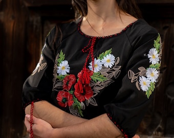May include: A woman wearing a black embroidered blouse with red and white floral designs. The blouse has red tassels on the sleeves and neckline.