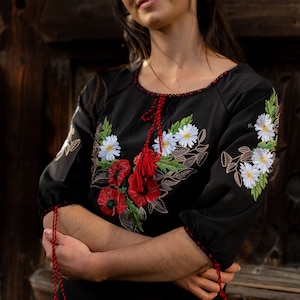 May include: A woman wearing a black embroidered blouse with red and white floral designs. The blouse has red tassels on the sleeves and neckline.