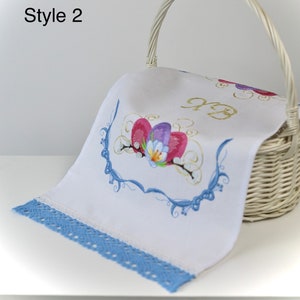 May include: A white woven basket holds a white cloth with embroidered flowers in pink, purple, and blue. The cloth has a blue lace trim. The text "Style 2" is in the upper left corner.