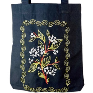 May include: Black canvas tote bag with a floral design embroidered in yellow and white. The design is framed by a border of yellow leaves.