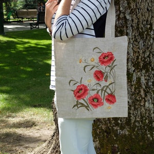 Op de afbeelding: Een linnen tas met geborduurde rode klaprozen en witte madeliefjes. De tas heeft een natuurlijke linnen kleur en wordt vastgehouden door iemand die een gestreept shirt en een witte broek draagt.