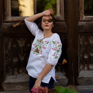 May include: A woman with long brown hair wearing a white embroidered blouse with red and green floral designs and blue jeans. She is standing in front of a wooden door with a window.