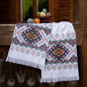 May include: A white embroidered table runner with a colorful geometric pattern. The runner is hanging on a wooden surface with a bowl of painted Easter eggs in the background.