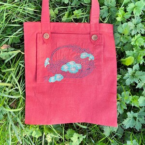 Op de afbeelding: Een rode tote bag met een geborduurd bloemenontwerp op de voorzak. De tas heeft twee bruine knopen aan de voorkant.
